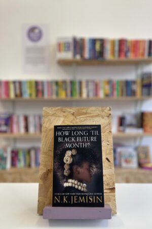 Cymraeg: Copi o 'How Long 'Til Black Future Month?' yn sefyll ar stondin llyfrau, tu blaen silffoedd o lyfrau yn y cefndir. English: A copy of 'How Long 'Til Black Future Month?' sits on a stand in front of multiple shelves of other books.