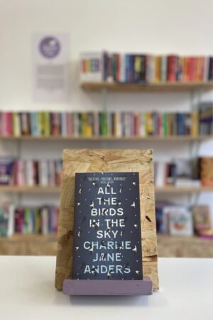 A copy of 'All the Birds in the Sky' sits on a stand in front of multiple shelves of other books.