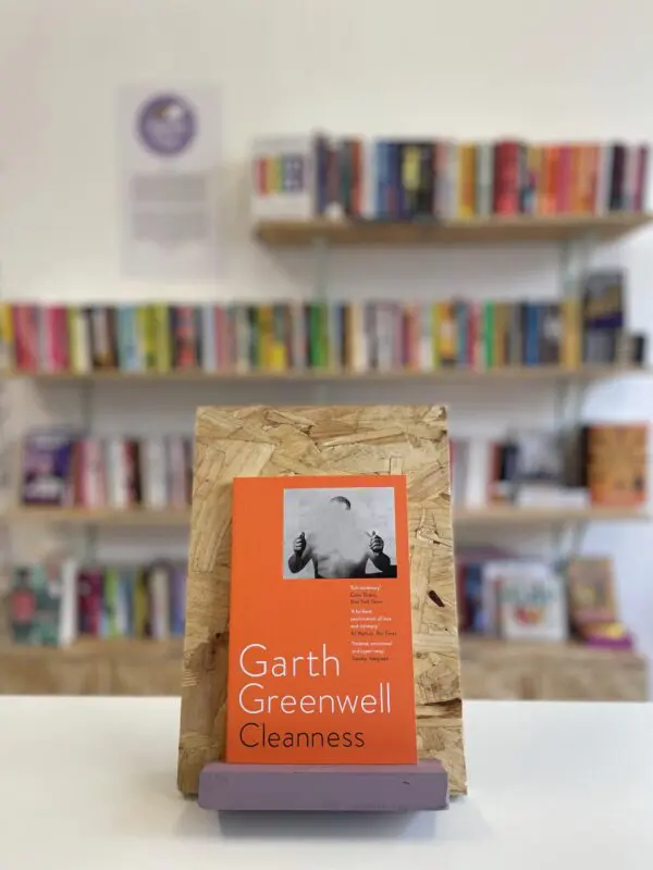 Cymraeg: Copi o 'Cleanness' yn sefyll ar stondin llyfrau, tu blaen silffoedd o lyfrau yn y cefndir. | English: A copy of 'Cleannes' sits on a stand in front of multiple shelves of other books.