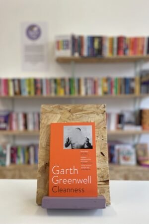 Cymraeg: Copi o 'Cleanness' yn sefyll ar stondin llyfrau, tu blaen silffoedd o lyfrau yn y cefndir. | English: A copy of 'Cleannes' sits on a stand in front of multiple shelves of other books.