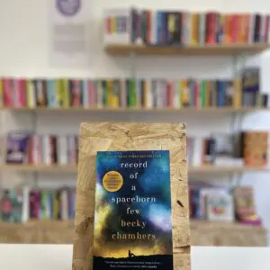 Cymraeg: Copi o 'Record of a Spaceborn Few' yn sefyll ar stondin llyfrau, tu blaen silffoedd o lyfrau yn y cefndir. English: A copy of 'Record of a Spaceborn Few' sits on a stand in front of multiple shelves of other books.