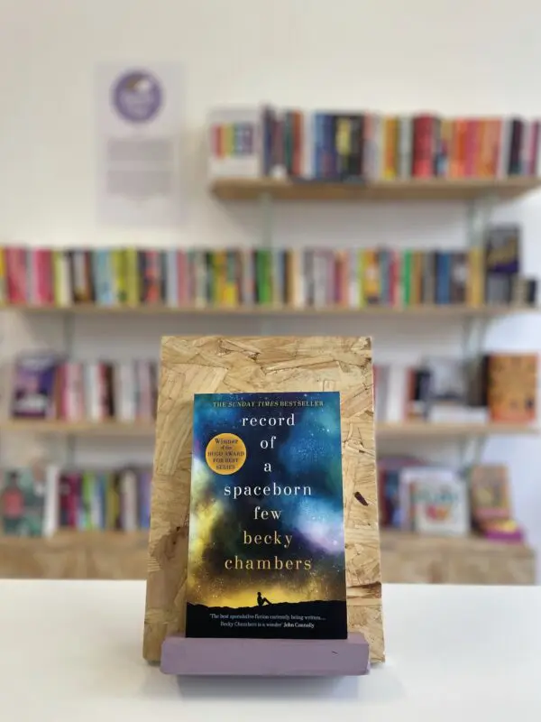Cymraeg: Copi o 'Record of a Spaceborn Few' yn sefyll ar stondin llyfrau, tu blaen silffoedd o lyfrau yn y cefndir. English: A copy of 'Record of a Spaceborn Few' sits on a stand in front of multiple shelves of other books.