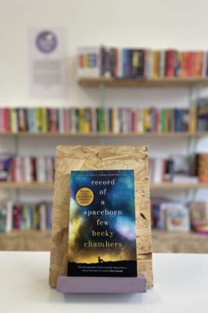 Cymraeg: Copi o 'Record of a Spaceborn Few' yn sefyll ar stondin llyfrau, tu blaen silffoedd o lyfrau yn y cefndir. English: A copy of 'Record of a Spaceborn Few' sits on a stand in front of multiple shelves of other books.