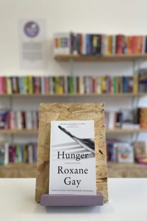 Cymraeg: Copi o 'Hunger: A Memoir of (My) Body' yn sefyll ar stondin llyfrau, tu blaen silffoedd o lyfrau yn y cefndir. | English: A copy of 'Hunger: A Memoir of (My) Body' sits on a stand in front of multiple shelves of other books.