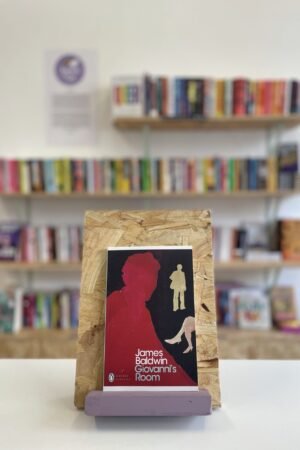 Cymraeg: Copi o 'Giovanni's Room' yn sefyll ar stondin llyfrau, tu blaen silffoedd o lyfrau yn y cefndir. | English: A copy of 'Giovanni's Room' sits on a stand in front of multiple shelves of other books.