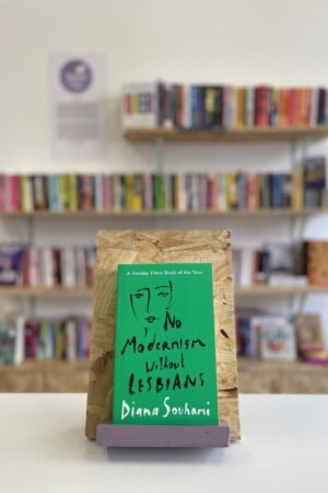 Cymraeg: Copi o 'No Modernism Without Lesbians' yn sefyll ar stondin llyfrau, tu blaen silffoedd o lyfrau yn y cefndir. English: A copy of 'No Modernism Without Lesbians' sits on a stand in front of multiple shelves of other books.