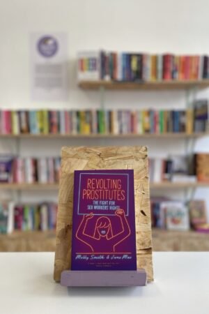 Cymraeg: Copi o 'Revolting Prostitutes' yn sefyll ar stondin llyfrau, tu blaen silffoedd o lyfrau yn y cefndir. | English: A copy of 'Revolting Prostitutes' sits on a stand in front of multiple shelves of other books.
