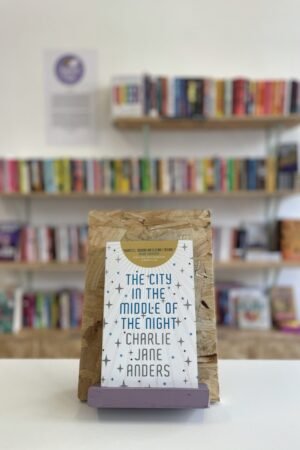 Cymraeg: Copi o 'The City in the Middle of the Night' yn sefyll ar stondin llyfrau, tu blaen silffoedd o lyfrau yn y cefndir. | English: A copy of 'The City in the Middle of the Night' sits on a stand in front of multiple shelves of other books.