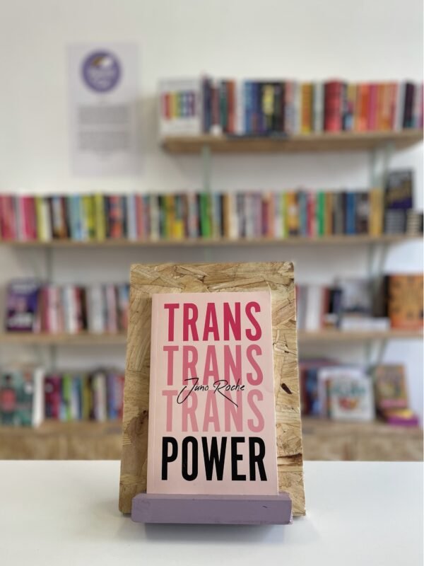 Cymraeg: Copi o 'Trans Power' yn sefyll ar stondin llyfrau, tu blaen silffoedd o lyfrau yn y cefndir. English: A copy of 'Trans Power' sits on a stand in front of multiple shelves of other books.