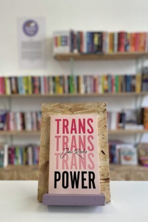 Cymraeg: Copi o 'Trans Power' yn sefyll ar stondin llyfrau, tu blaen silffoedd o lyfrau yn y cefndir. English: A copy of 'Trans Power' sits on a stand in front of multiple shelves of other books.