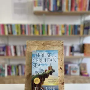 Cymraeg: Copi o 'The House in the Cerulean Sea' yn sefyll ar stondin llyfrau, tu blaen silffoedd o lyfrau yn y cefndir. | English: A copy of 'The House in the Cerulean Sea' sits on a stand in front of multiple shelves of other books.