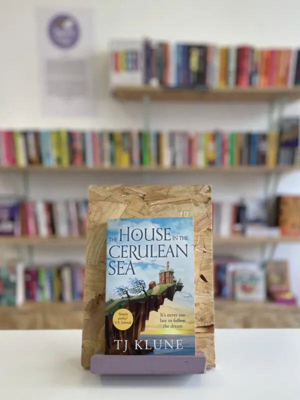 Cymraeg: Copi o 'The House in the Cerulean Sea' yn sefyll ar stondin llyfrau, tu blaen silffoedd o lyfrau yn y cefndir. | English: A copy of 'The House in the Cerulean Sea' sits on a stand in front of multiple shelves of other books.