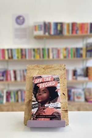 Cymraeg: Copi o 'Off the Record' yn sefyll ar stondin llyfrau, tu blaen silffoedd o lyfrau yn y cefndir. | English: A copy of 'Off the Record' sits on a stand in front of multiple shelves of other books.