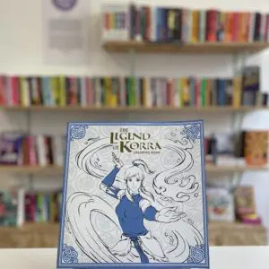 Cymraeg: Copi o 'The Legend of Korra Coloring Book' yn sefyll ar stondin llyfrau, tu blaen silffoedd o lyfrau yn y cefndir. English: A copy of 'The Legend of Korra Coloring Book' sits on a stand in front of multiple shelves of other books.