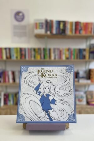 Cymraeg: Copi o 'The Legend of Korra Coloring Book' yn sefyll ar stondin llyfrau, tu blaen silffoedd o lyfrau yn y cefndir. English: A copy of 'The Legend of Korra Coloring Book' sits on a stand in front of multiple shelves of other books.