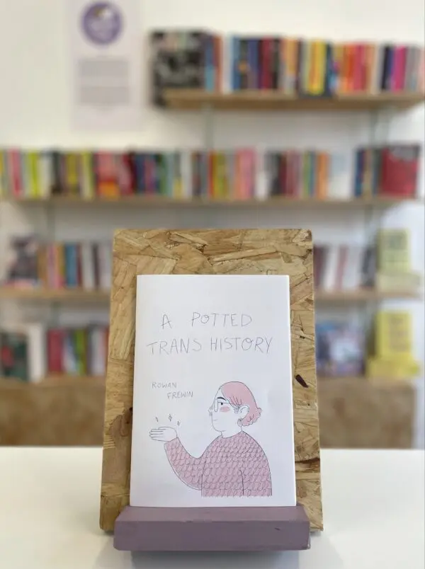 A copy of 'A Potted Trans History' sits on a stand in front of multiple shelves of other books.