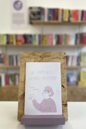 A copy of 'A Potted Trans History' sits on a stand in front of multiple shelves of other books.