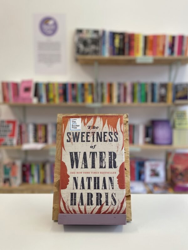 Cymraeg: Copi o 'The Sweetness of Water' yn sefyll ar stondin llyfrau, tu blaen silffoedd o lyfrau yn y cefndir. | English: A copy of 'The Sweetness of Water' sits on a stand in front of multiple shelves of other books.