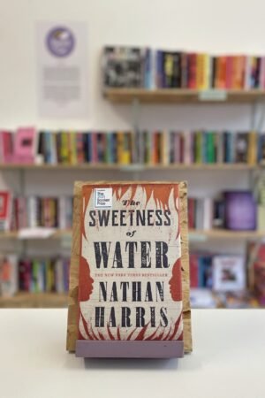 Cymraeg: Copi o 'The Sweetness of Water' yn sefyll ar stondin llyfrau, tu blaen silffoedd o lyfrau yn y cefndir. | English: A copy of 'The Sweetness of Water' sits on a stand in front of multiple shelves of other books.
