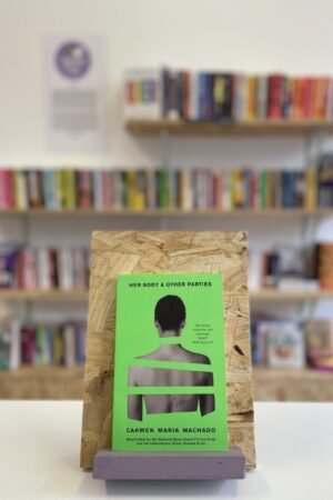 A copy of 'Her Body & Other Parties' sits on a stand in front of multiple shelves of other books.
