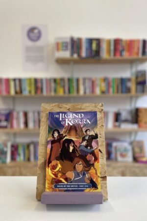 Cymraeg: Copi o 'The Legend of Korra: Ruins of the Empire, Part One' yn sefyll ar stondin llyfrau, tu blaen silffoedd o lyfrau yn y cefndir. | English: A copy of 'The Legend of Korra: Ruins of the Empire, Part One' sits on a stand in front of multiple shelves of other books.