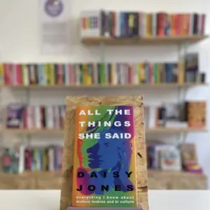 Cymraeg: Copi o 'All the Things She Said' yn sefyll ar stondin llyfrau, tu blaen silffoedd o lyfrau yn y cefndir. | English: A copy of 'All the Things She Said' sits on a stand in front of multiple shelves of other books.