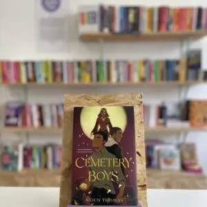 A copy of 'Cemetery Boys' sits on a stand in front of multiple shelves of other books.