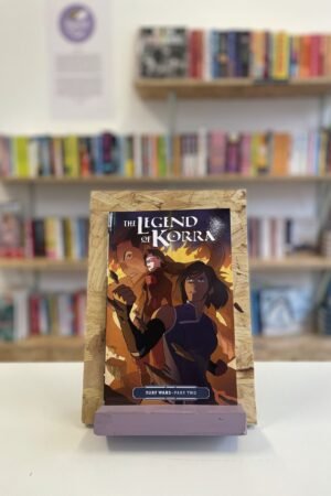 Cymraeg: Copi o 'The Legend of Korra: Turf Wars, Part Two' yn sefyll ar stondin llyfrau, tu blaen silffoedd o lyfrau yn y cefndir. | English: A copy of 'The Legend of Korra: Turf Wars, Part Two' sits on a stand in front of multiple shelves of other books.