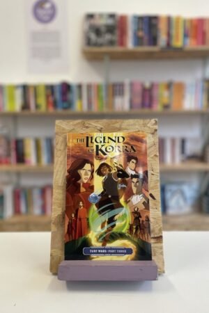 Cymraeg: Copi o 'The Legend of Korra: Turf Wars, Part Three' yn sefyll ar stondin llyfrau, tu blaen silffoedd o lyfrau yn y cefndir. | English: A copy of 'The Legend of Korra: Turf Wars, Part Three' sits on a stand in front of multiple shelves of other books.
