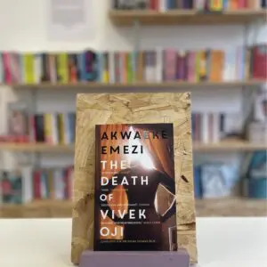 Cymraeg: Copi o 'The Death of Vivek Oji' yn sefyll ar stondin llyfrau, tu blaen silffoedd o lyfrau yn y cefndir. | English: A copy of 'The Death of Vivek Oji' sits on a stand in front of multiple shelves of other books.