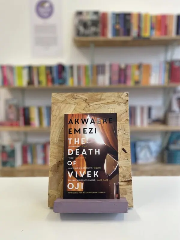 Cymraeg: Copi o 'The Death of Vivek Oji' yn sefyll ar stondin llyfrau, tu blaen silffoedd o lyfrau yn y cefndir. | English: A copy of 'The Death of Vivek Oji' sits on a stand in front of multiple shelves of other books.