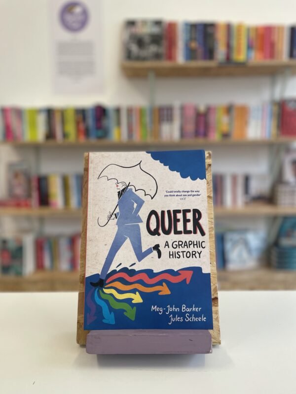 Cymraeg: Copi o 'Queer: A Graphic History' yn sefyll ar stondin llyfrau, tu blaen silffoedd o lyfrau yn y cefndir. | English: A copy of 'Queer: A Graphic History' sits on a stand in front of multiple shelves of other books.
