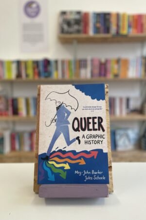 Cymraeg: Copi o 'Queer: A Graphic History' yn sefyll ar stondin llyfrau, tu blaen silffoedd o lyfrau yn y cefndir. | English: A copy of 'Queer: A Graphic History' sits on a stand in front of multiple shelves of other books.