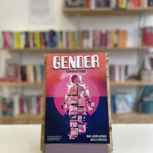 Cymraeg: Copi o 'Gender: A Graphic Guide' yn sefyll ar stondin llyfrau, tu blaen silffoedd o lyfrau yn y cefndir. | English: A copy of 'Gender: A Graphic Guide' sits on a stand in front of multiple shelves of other books.