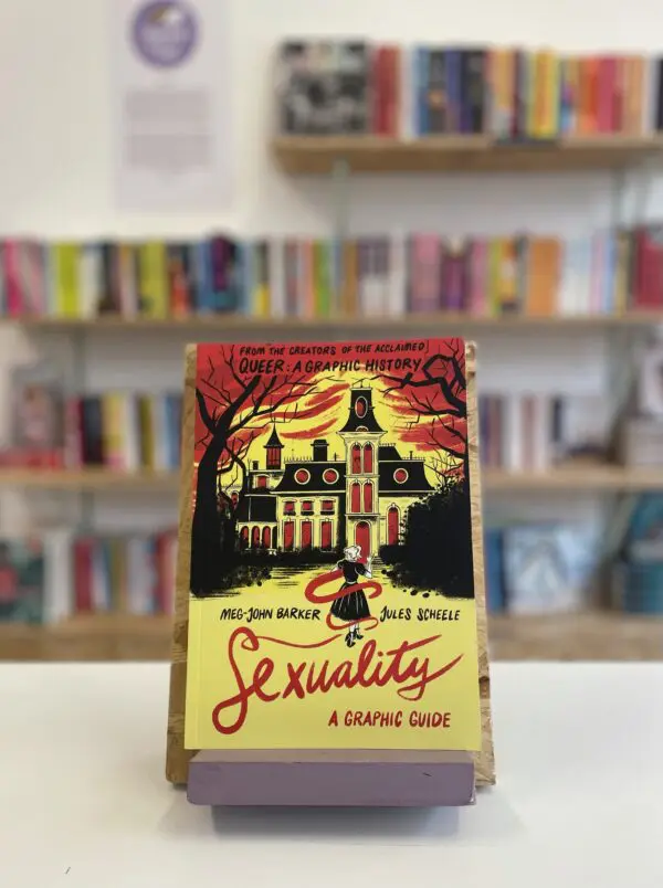 Cymraeg: Copi o 'Sexuality: A Graphic Guide' yn sefyll ar stondin llyfrau, tu blaen silffoedd o lyfrau yn y cefndir. | English: A copy of 'Sexuality: A Graphic Guide' sits on a stand in front of multiple shelves of other books.