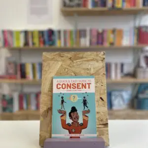 Cymraeg: Copi o 'A Quick & Easy Guide to Consent' yn sefyll ar stondin llyfrau, tu blaen silffoedd o lyfrau yn y cefndir. | English: A copy of 'A Quick & Easy Guide to Consent' sits on a stand in front of multiple shelves of other books.