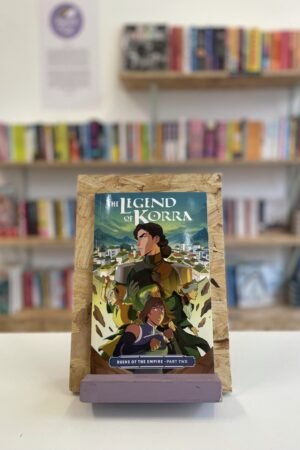 Cymraeg: Copi o 'The Legend of Korra: Ruins of the Empire, Part Two' yn sefyll ar stondin llyfrau, tu blaen silffoedd o lyfrau yn y cefndir. | English: A copy of 'The Legend of Korra: Ruins of the Empire, Part Two' sits on a stand in front of multiple shelves of other books.