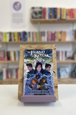 Cymraeg: Copi o 'The Legend of Korra: Ruins of the Empire, Part Three' yn sefyll ar stondin llyfrau, tu blaen silffoedd o lyfrau yn y cefndir. | English: A copy of 'The Legend of Korra: Ruins of the Empire, Part Three' sits on a stand in front of multiple shelves of other books.