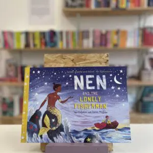 Cymraeg: Copi o 'Nen and the Lonely Fisherman' yn sefyll ar stondin llyfrau, tu blaen silffoedd o lyfrau yn y cefndir. | English: A copy of 'Nen and the Lonely Fisherman' sits on a stand in front of multiple shelves of other books.