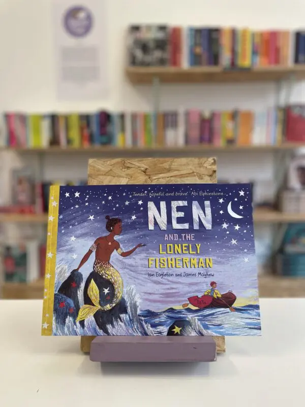 Cymraeg: Copi o 'Nen and the Lonely Fisherman' yn sefyll ar stondin llyfrau, tu blaen silffoedd o lyfrau yn y cefndir. | English: A copy of 'Nen and the Lonely Fisherman' sits on a stand in front of multiple shelves of other books.