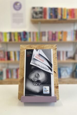 Cymraeg: Copi o 'The Promise yn sefyll ar stondin llyfrau, tu blaen silffoedd o lyfrau yn y cefndir. | English: A copy of 'The Promise' sits on a stand in front of multiple shelves of other books.