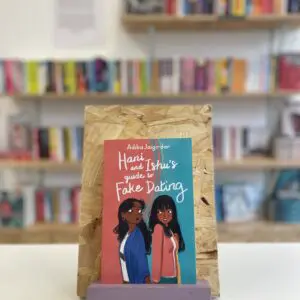 Cymraeg: Copi o 'Hani and Ishu's Guide to Fake Dating' yn sefyll ar stondin llyfrau, tu blaen silffoedd o lyfrau yn y cefndir. | English: A copy of 'Hani and Ishu's Guide to Fake Dating' sits on a stand in front of multiple shelves of other books.