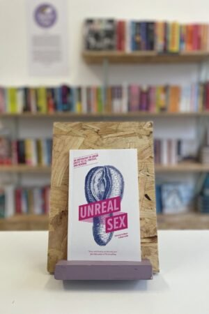 Cymraeg: Copi o 'Unreal Sex' yn sefyll ar stondin llyfrau, tu blaen silffoedd o lyfrau yn y cefndir. | English: A copy of 'Unreal Sex' sits on a stand in front of multiple shelves of other books.