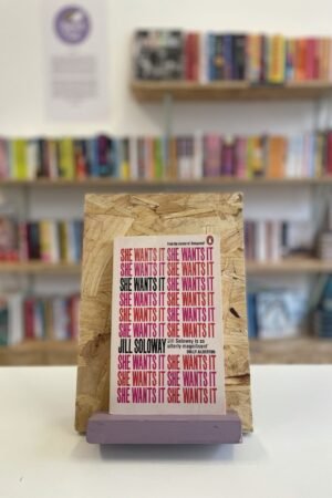 Cymraeg: Copi o 'She Wants It' yn sefyll ar stondin llyfrau, tu blaen silffoedd o lyfrau yn y cefndir. | English: A copy of 'She Wants It' sits on a stand in front of multiple shelves of other books.