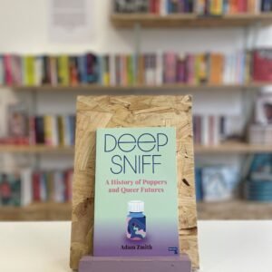 Cymraeg: Copi o 'Deep Sniff' yn sefyll ar stondin llyfrau, tu blaen silffoedd o lyfrau yn y cefndir. | English: A copy of 'Deep Sniff' sits on a stand in front of multiple shelves of other books.