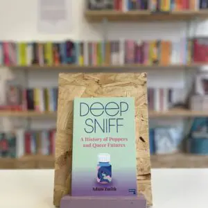 Cymraeg: Copi o 'Deep Sniff' yn sefyll ar stondin llyfrau, tu blaen silffoedd o lyfrau yn y cefndir. | English: A copy of 'Deep Sniff' sits on a stand in front of multiple shelves of other books.