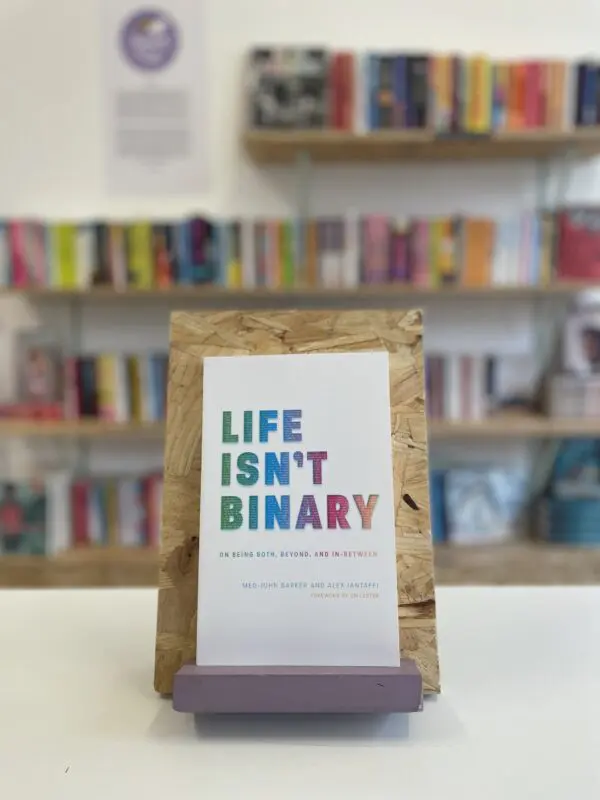 Cymraeg: Copi o 'Life Isn't Binary' yn sefyll ar stondin llyfrau, tu blaen silffoedd o lyfrau yn y cefndir. | English: A copy of 'Life Isn't Binary' sits on a stand in front of multiple shelves of other books.