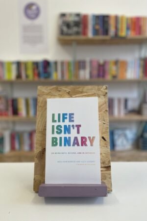 Cymraeg: Copi o 'Life Isn't Binary' yn sefyll ar stondin llyfrau, tu blaen silffoedd o lyfrau yn y cefndir. | English: A copy of 'Life Isn't Binary' sits on a stand in front of multiple shelves of other books.