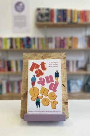 Cymraeg: Copi o 'Fat and Queer' yn sefyll ar stondin llyfrau, tu blaen silffoedd o lyfrau yn y cefndir. | English: A copy of 'Fat and Queer' sits on a stand in front of multiple shelves of other books.