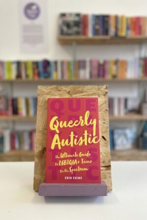 Cymraeg: Copi o 'Queerly Autistic' yn sefyll ar stondin llyfrau, tu blaen silffoedd o lyfrau yn y cefndir. | English: A copy of 'Queerly Autistic' sits on a stand in front of multiple shelves of other books.